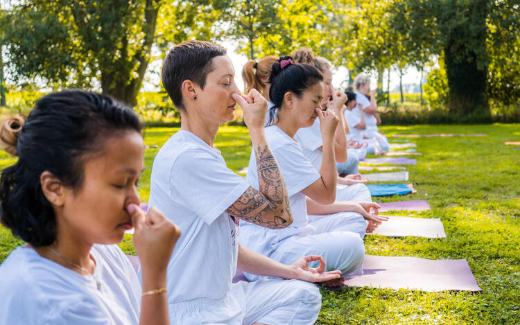 Arhanta Yoga students sit outside on a yoga mat and close their nostrils while practicing Ujjayi Pranayama